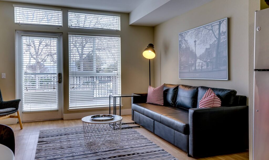 Modern living room with a black leather sofa, striped pillows, floor lamp, coffee table, and large window with blinds. A framed photo hangs on the wall above the sofa. Light wood flooring and a striped rug are visible.
