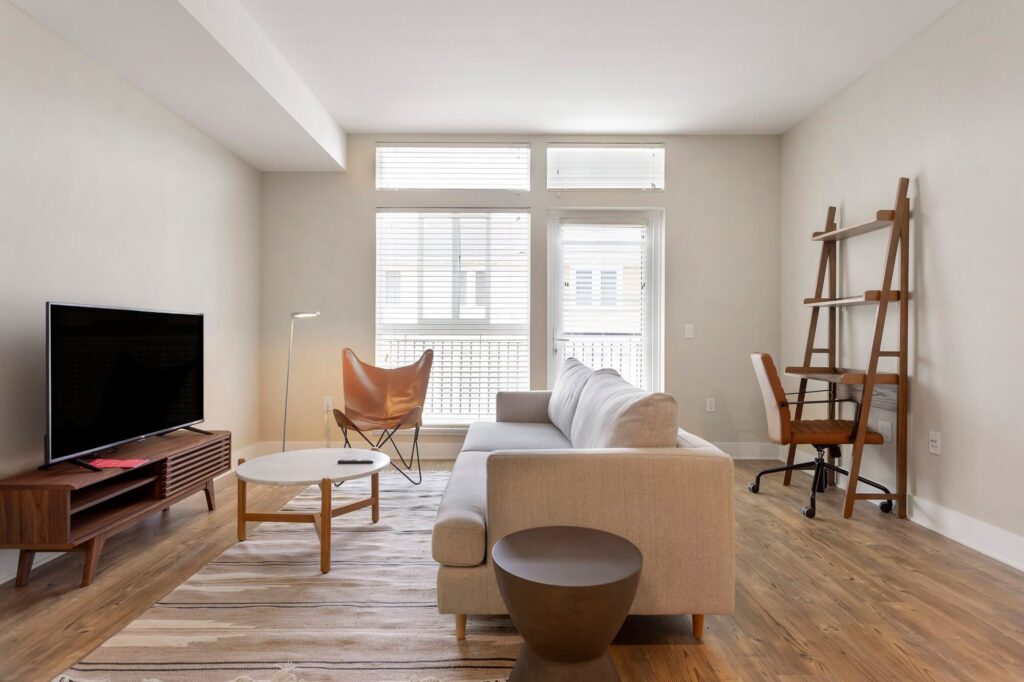 Modern living room with a beige sofa, TV on a wooden stand, butterfly chair, small round table, rug, lamp, and ladder-style desk with a chair. Large windows let in natural light.