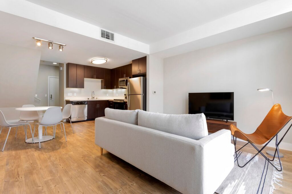 Modern apartment with a gray sofa, tan leather accent chair, TV, round dining table with four white chairs, wooden flooring, and an open kitchen with dark cabinets and stainless steel appliances.