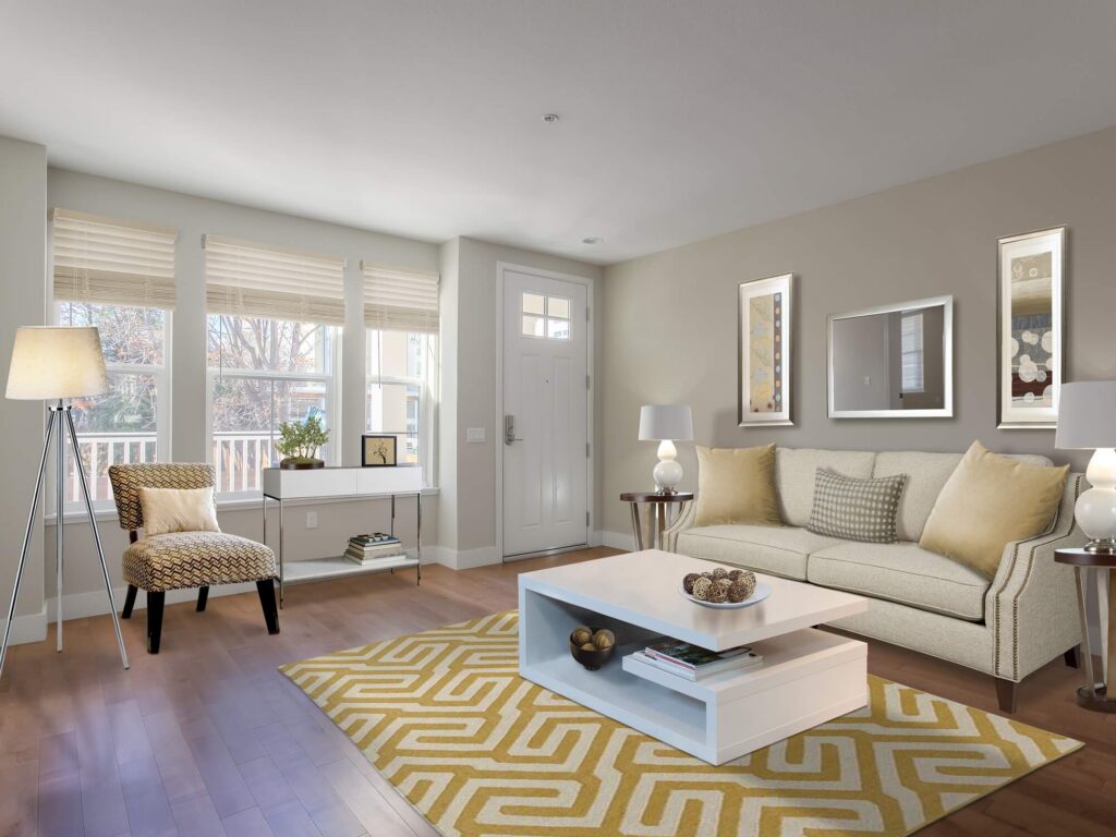 Modern living room with beige sofa, yellow patterned rug, white coffee table, accent chair, lamps, wall art, and large windows letting in natural light. A small table with a plant sits by the front door.