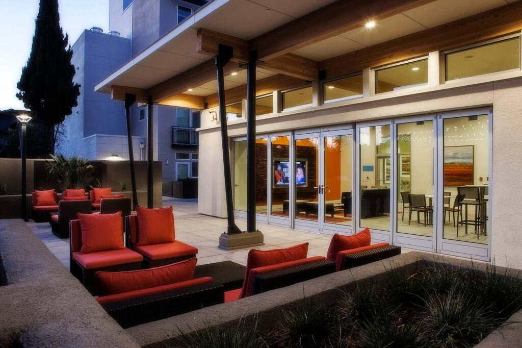 Modern outdoor patio with red cushioned seating and tables facing large glass doors. Inside, a TV, pool table, and dining area are visible. Overhead lights illuminate the cozy, contemporary space at dusk.