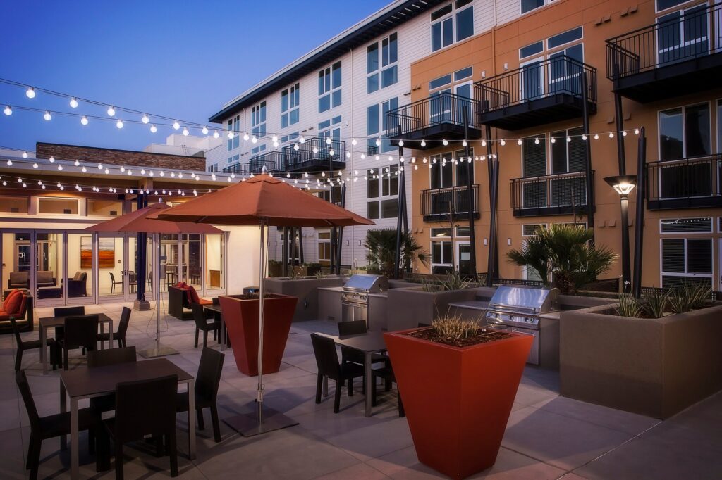 Modern apartment courtyard at dusk with string lights, tables, chairs, red umbrellas, large planters, and outdoor grills. Multi-story buildings with balconies surround the well-lit patio area.