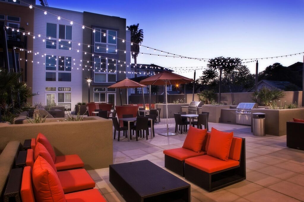 Outdoor patio with red-cushioned seating, tables, umbrellas, string lights overhead, and built-in grills. Modern apartment building in the background at dusk. Relaxing and inviting atmosphere.