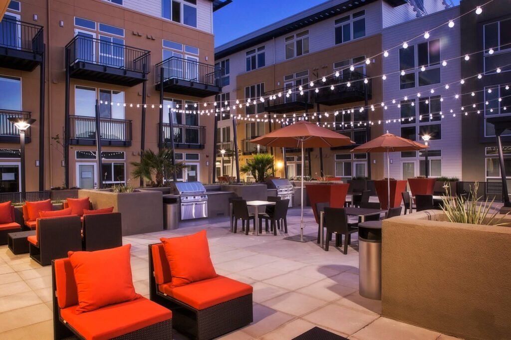 Modern apartment courtyard at dusk with string lights, red-cushioned lounge chairs, dining tables with umbrellas, and an outdoor grill area, surrounded by multi-story apartments with balconies.
