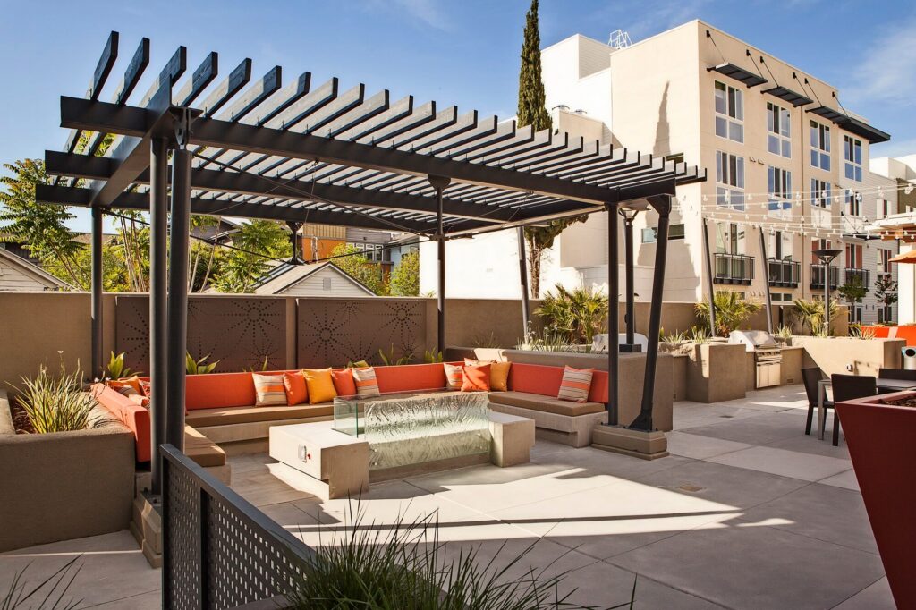Modern rooftop patio with a black pergola, orange and red cushioned seating, a glass fire pit, planters, and apartment buildings in the background under a blue sky.