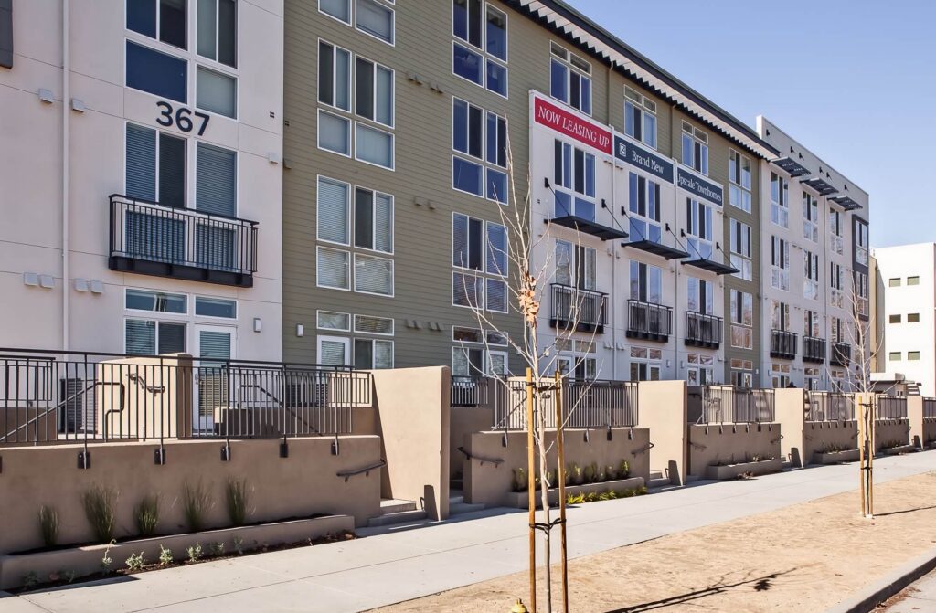 Modern apartment building with large windows and balconies, a Now Leasing sign, a sidewalk, young trees, and clear blue sky.