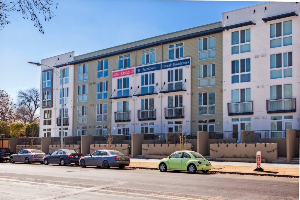 A modern apartment building features Now Leasing and other banners. Several cars, including a green Volkswagen Beetle, are parked along the street in front of the building.