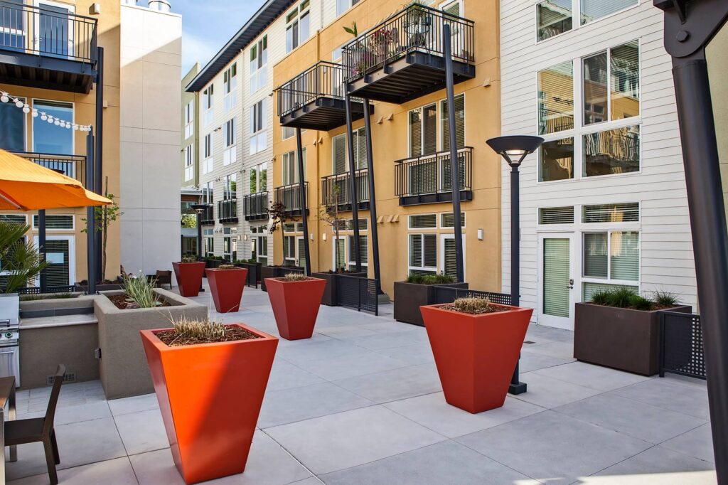 Modern apartment courtyard with tall orange planters, outdoor seating, balconies, and large windows, surrounded by multi-story cream and white buildings under a blue sky.