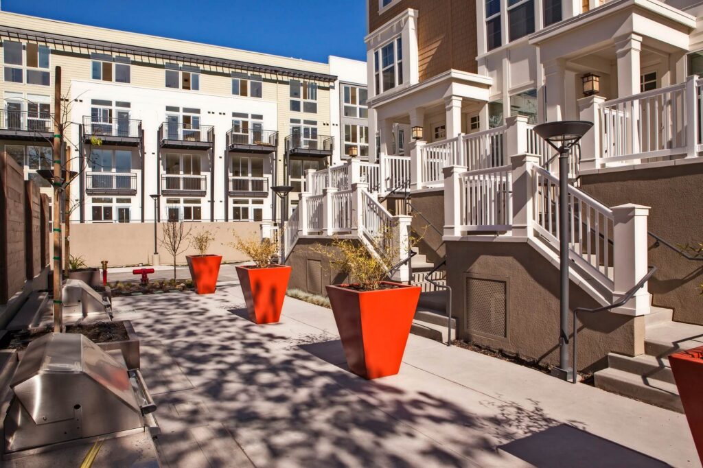 Modern apartment complex courtyard with large orange planters, white stair railings, beige walls, and balconies. Benches line the left side; shadows from trees are visible on the ground. Blue sky above.