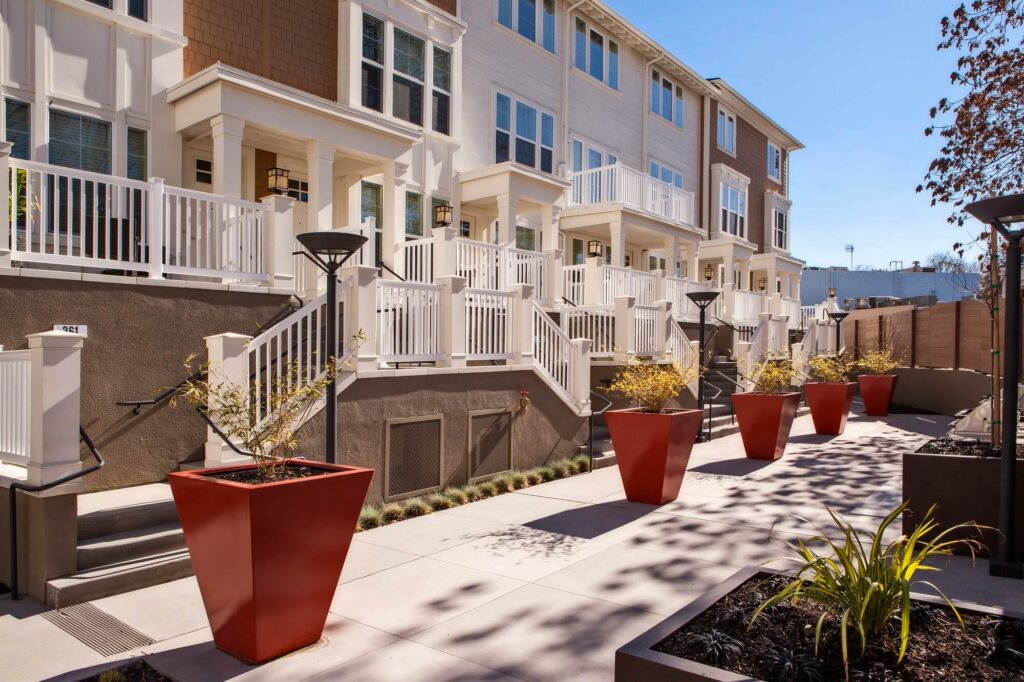 A row of modern townhouses with white railings and balconies, facing a walkway lined with large red planters and small shrubs on a sunny day.