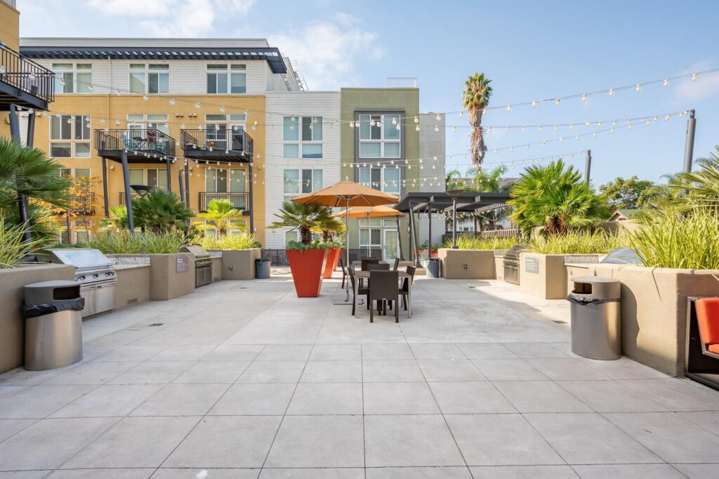 Modern apartment courtyard with BBQ grills, patio tables, orange umbrellas, string lights, and tall planters, surrounded by greenery and multi-story buildings under a clear sky.