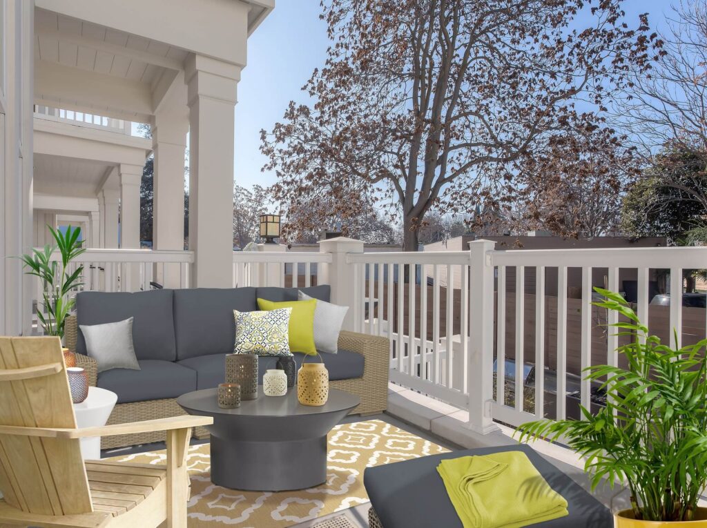 A cozy balcony with a gray outdoor sofa, yellow and white pillows, a round table, accent chairs, green plants, and a view of trees and houses under a clear blue sky.