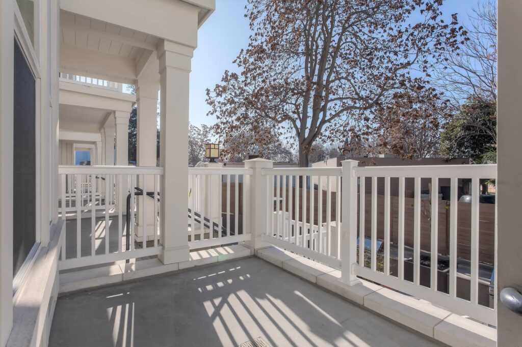 A spacious, sunlit balcony with white railings and columns, overlooking a street and large trees with sparse brown leaves under a clear blue sky.