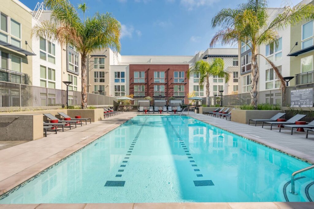Rectangular outdoor swimming pool with lane markings, surrounded by lounge chairs and palm trees, set in a modern apartment complex courtyard under a clear, sunny sky.