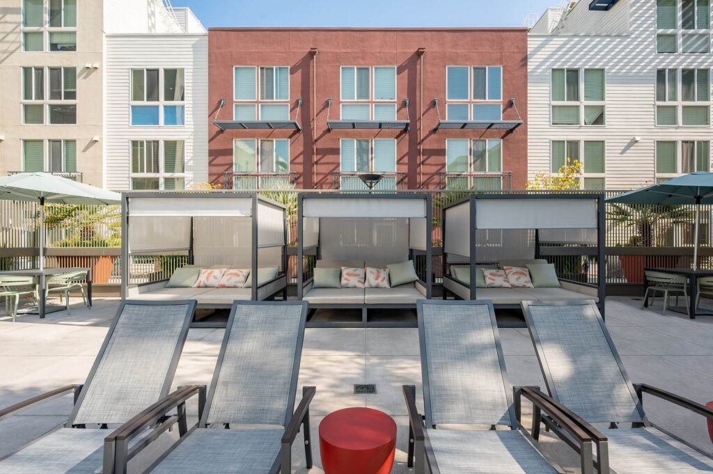 Four lounge chairs face poolside cabanas with cushions and pillows. Behind, modern apartment buildings feature large windows. There are patio tables, chairs, and umbrellas on a sunny day.