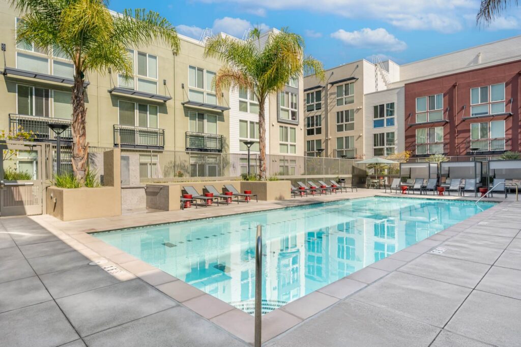 Modern apartment complex with a rectangular outdoor pool, lounge chairs, palm trees, and cabanas. The buildings have balconies and large windows, and the sky is partly cloudy.