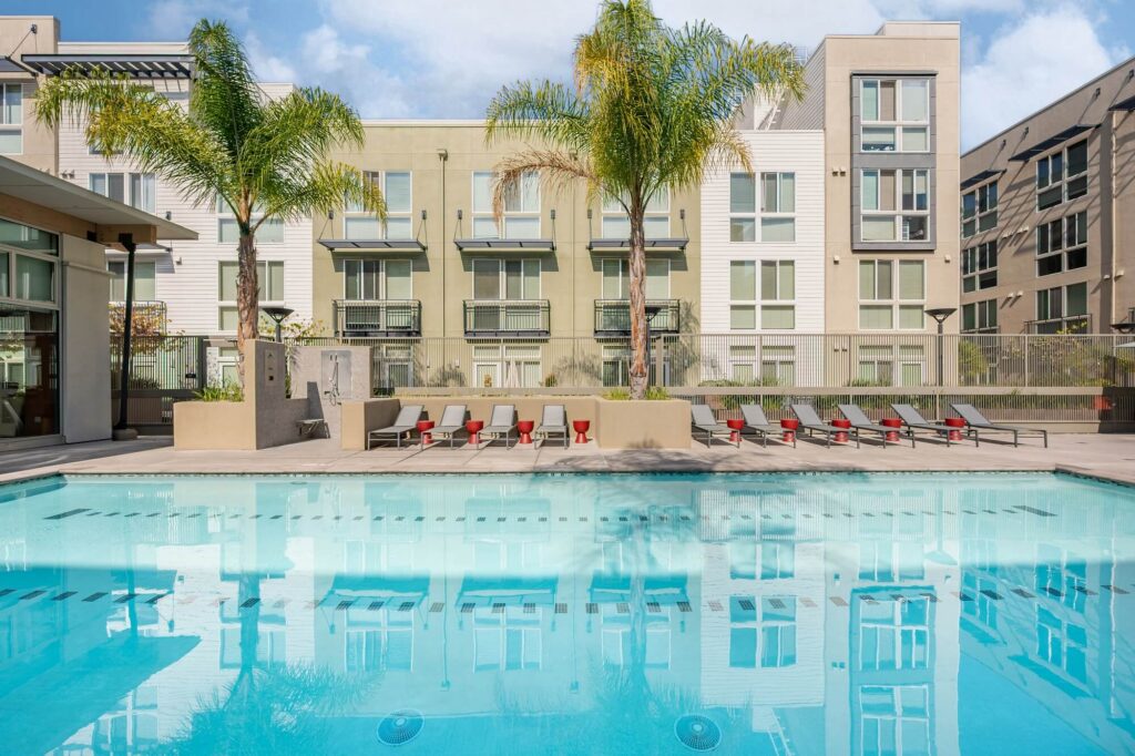 Modern apartment complex courtyard with a swimming pool, palm trees, lounge chairs, and several multi-story buildings in the background under a blue sky.