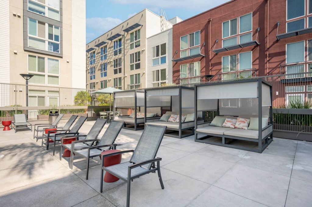 Outdoor lounge area with several gray lounge chairs, red side tables, and three shaded cabanas with cushioned seating, surrounded by modern apartment buildings. The space is open and well-lit.