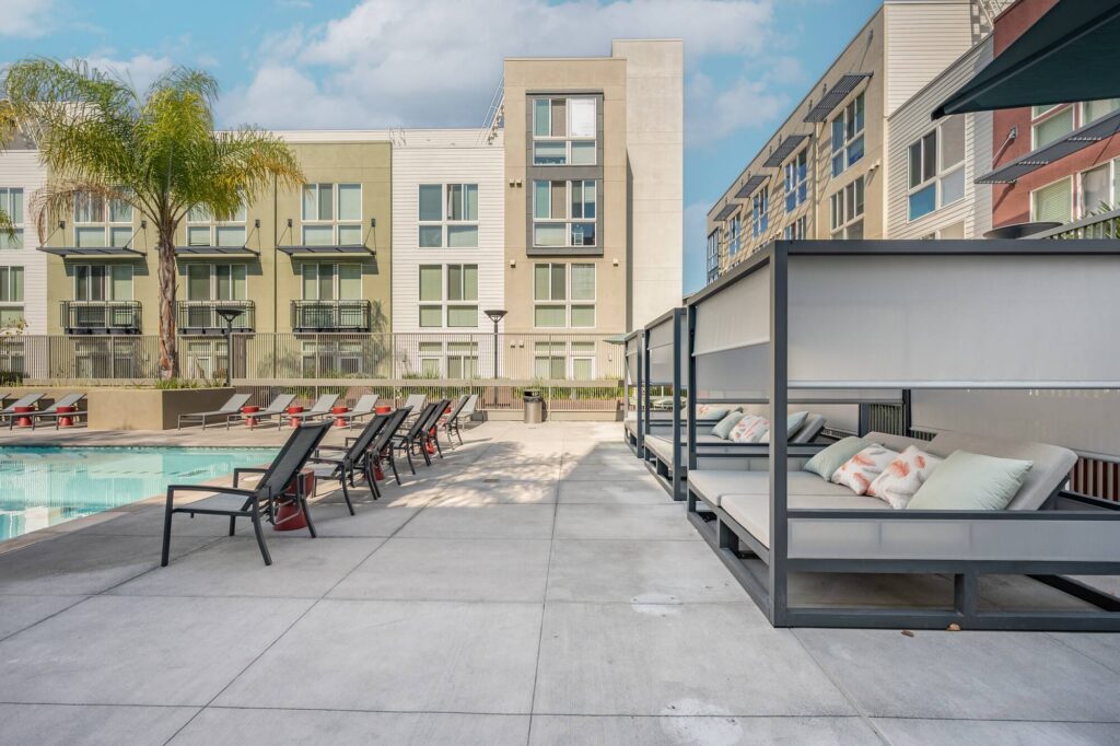 Modern apartment pool area with lounge chairs, shaded cabanas with pillows, and a palm tree. Several stories of apartment units are visible in the background under a partly cloudy sky.