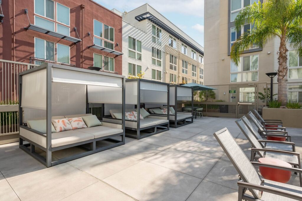 Outdoor lounge area with shaded cabanas containing cushioned seating and pillows, plus several reclining chairs nearby. Modern apartment buildings, palm tree, and blue sky are visible in the background.