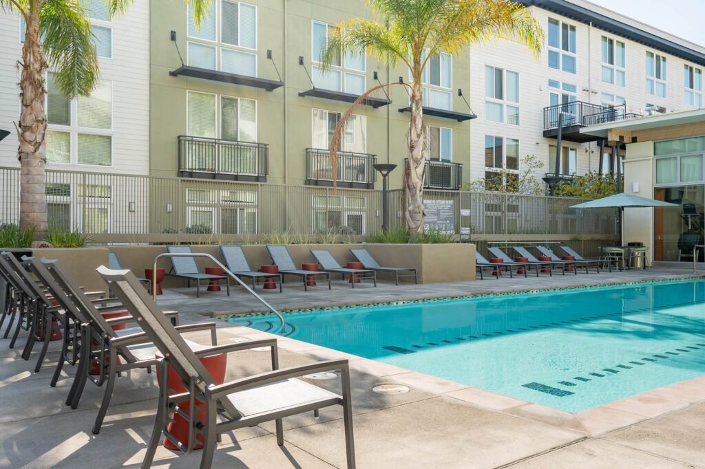 Modern apartment building with balconies, palm trees, and a swimming pool surrounded by lounge chairs and umbrellas on a sunny day.
