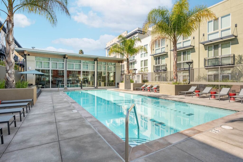 Outdoor pool surrounded by lounge chairs, palm trees, and modern apartment buildings under a partly cloudy sky.