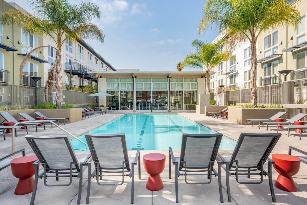 Modern outdoor swimming pool surrounded by lounge chairs, red side tables, tall palm trees, and apartment buildings under a blue sky.