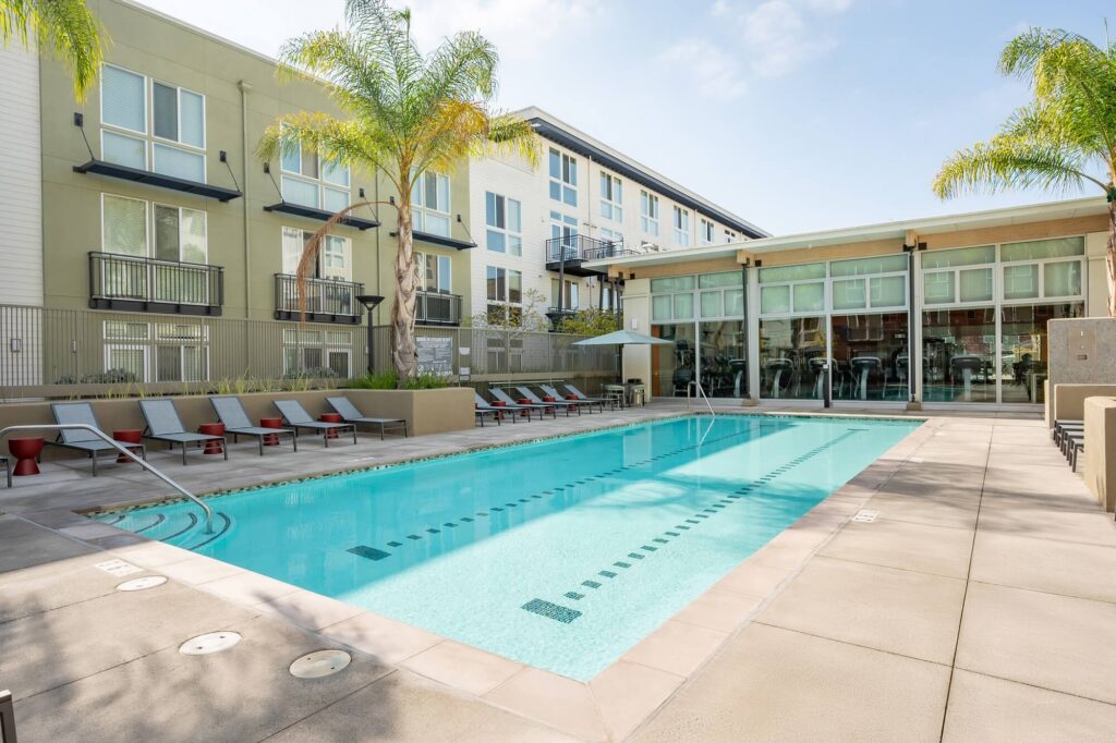 Outdoor swimming pool surrounded by lounge chairs and palm trees, with a modern apartment building and glass-walled clubhouse in the background on a sunny day.
